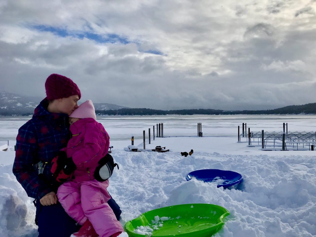 mother and daughter sitting in the snow with snow-covered lake ponderay in the background
