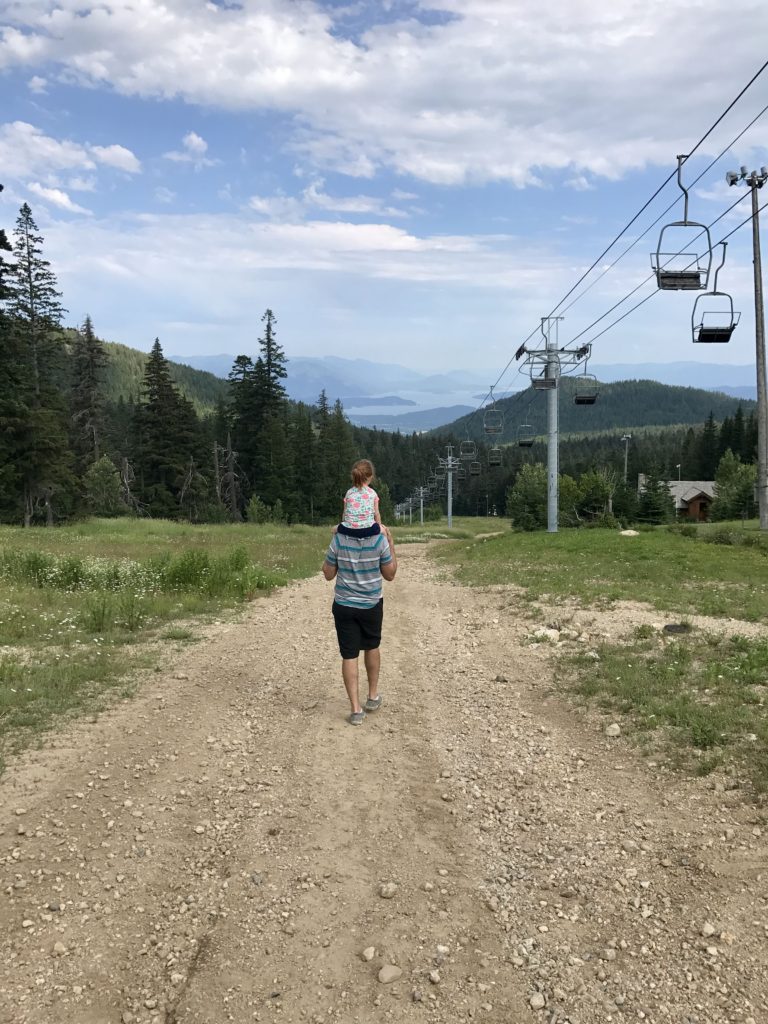 father walking down Schweitzer Mountain in the summer with a little girl on his shoulders.