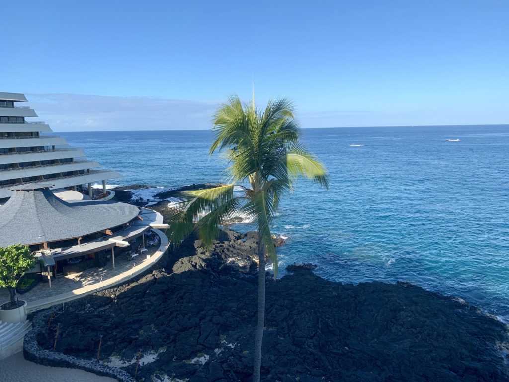 View of ocean, lava rock, palm tree and restaurant from hotel room balcony at Royal Kona Resort