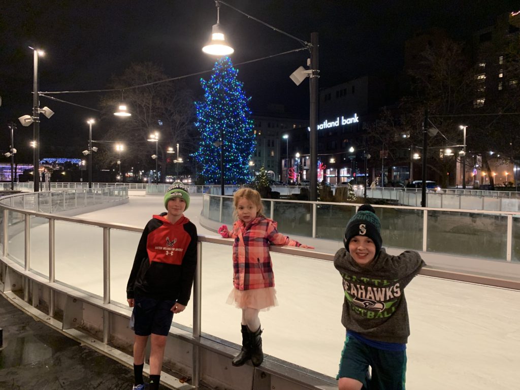 3 kids standing in front of Spokane Ice Ribbon in Riverfront Park with tree with blue lights on it in the background.