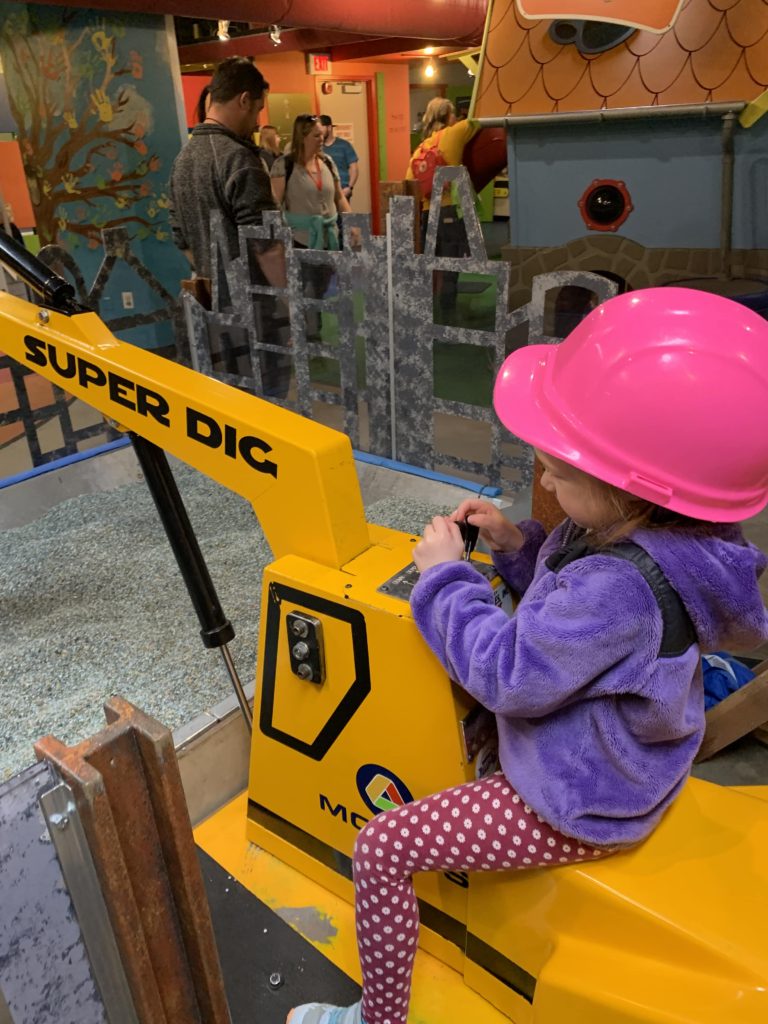 Child playing on fake construction equipment, wearing a hard hat, at Mobius Children's Museum